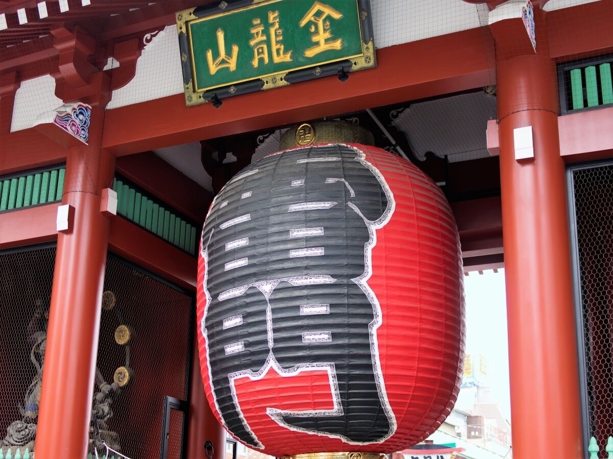 Un farol tradicional japonés colgado bajo una estructura de madera roja con un letrero verde encima.