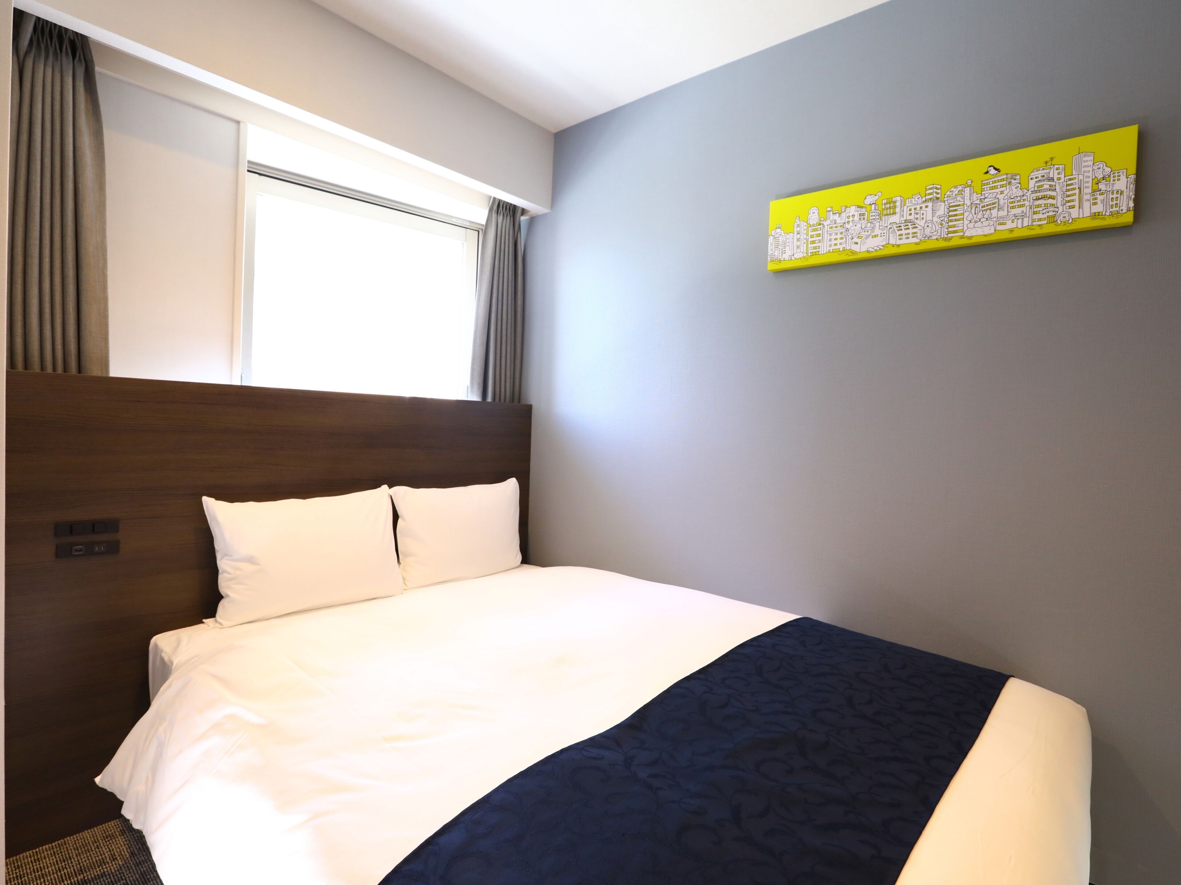 A hotel room with a bed, a window with curtains, a cityscape artwork on the wall, and a headboard.