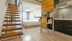 Una habitación de hotel moderna con una escalera de madera, una cama, un escritorio con una silla, una zona de cocina con armarios oscuros y un pequeño balcón.