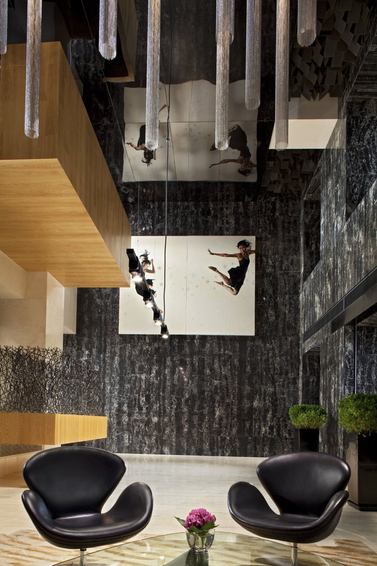 A modern lobby with a large mirror reflecting a person, black chairs, a wooden bench, and a vase with a flower.
