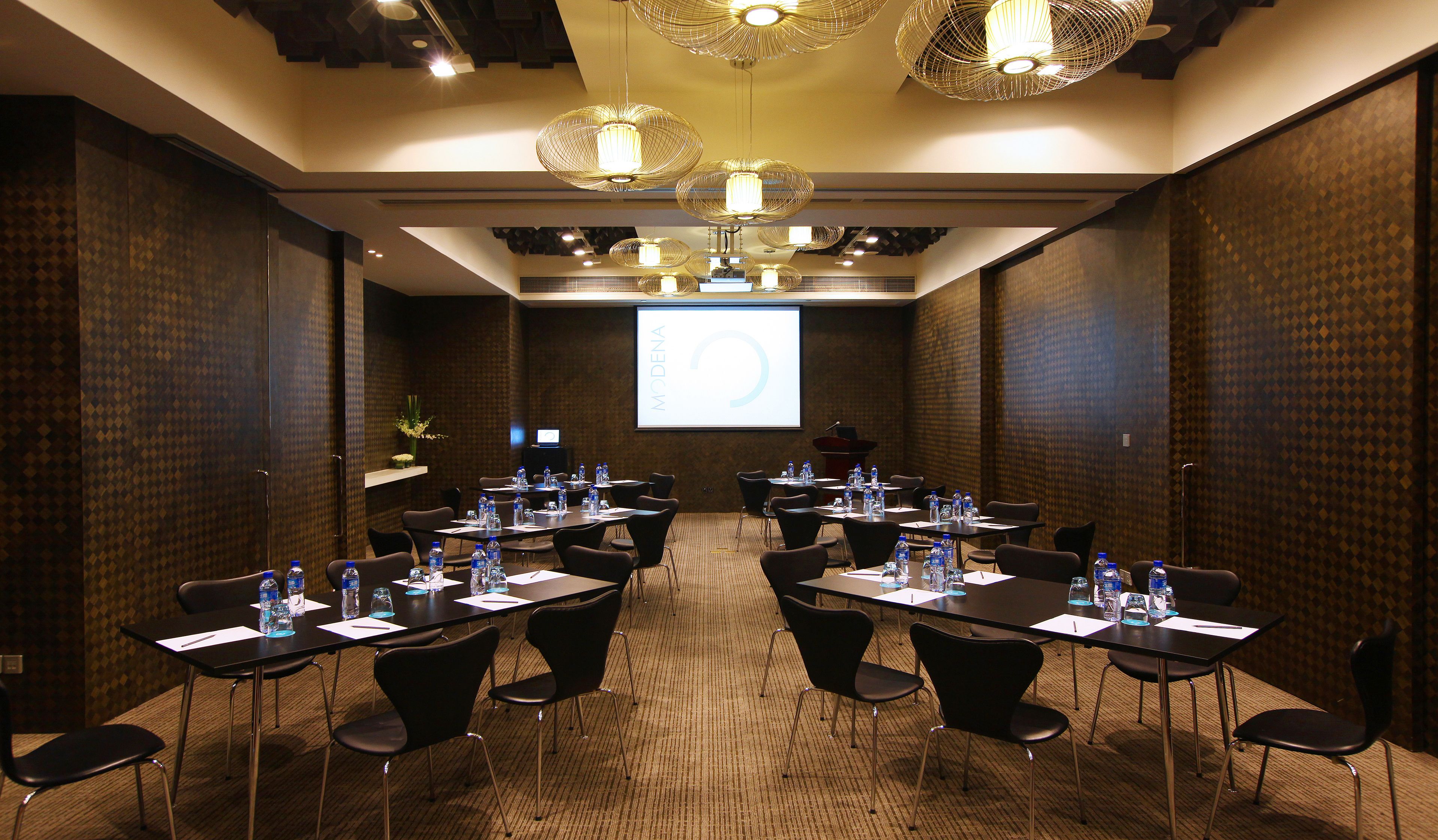 A conference room with a large screen, multiple tables, and chairs, equipped with water bottles and papers.