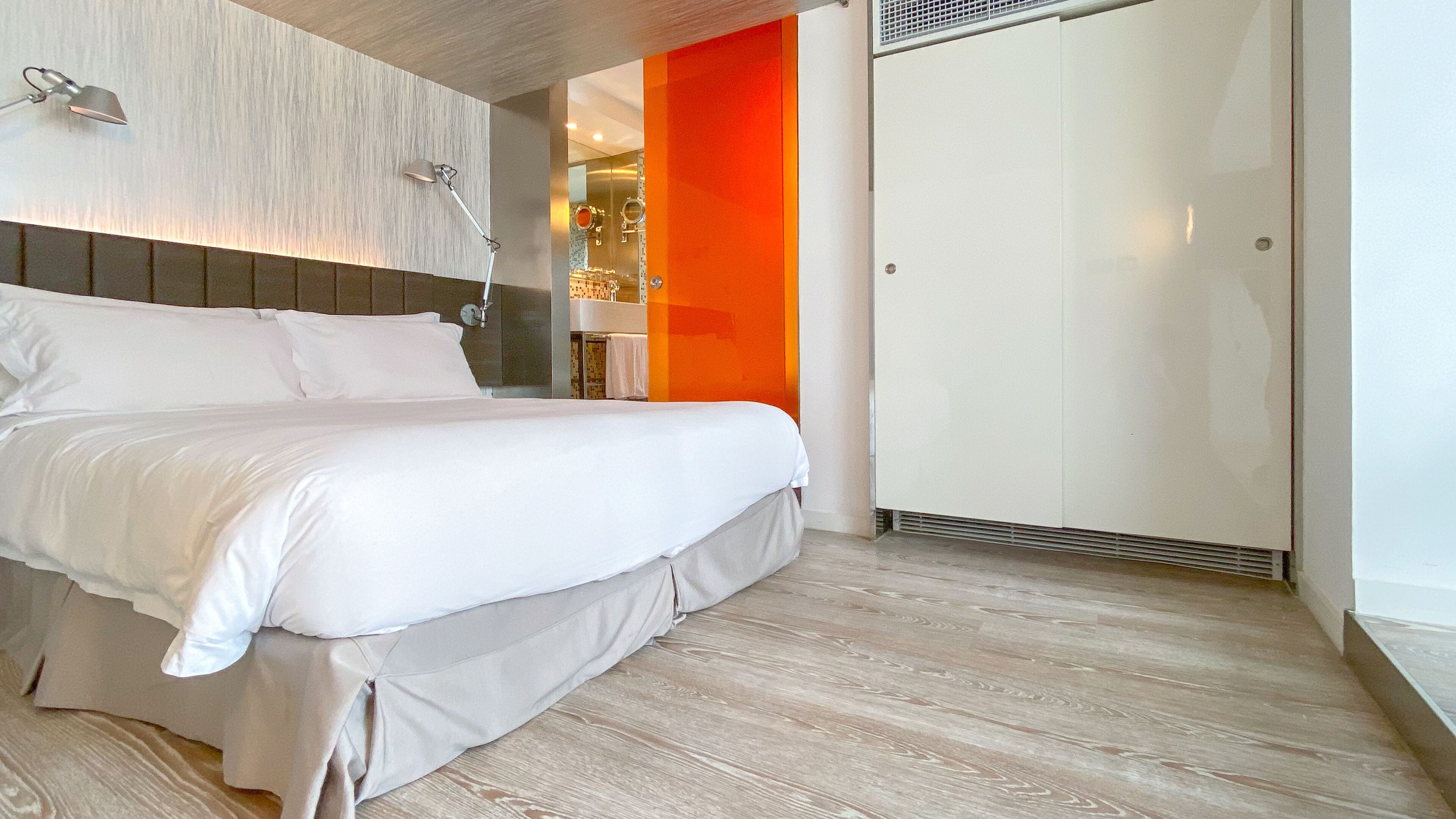 A modern hotel room with a large bed, a headboard with integrated lighting, a wardrobe, and a visible bathroom area.