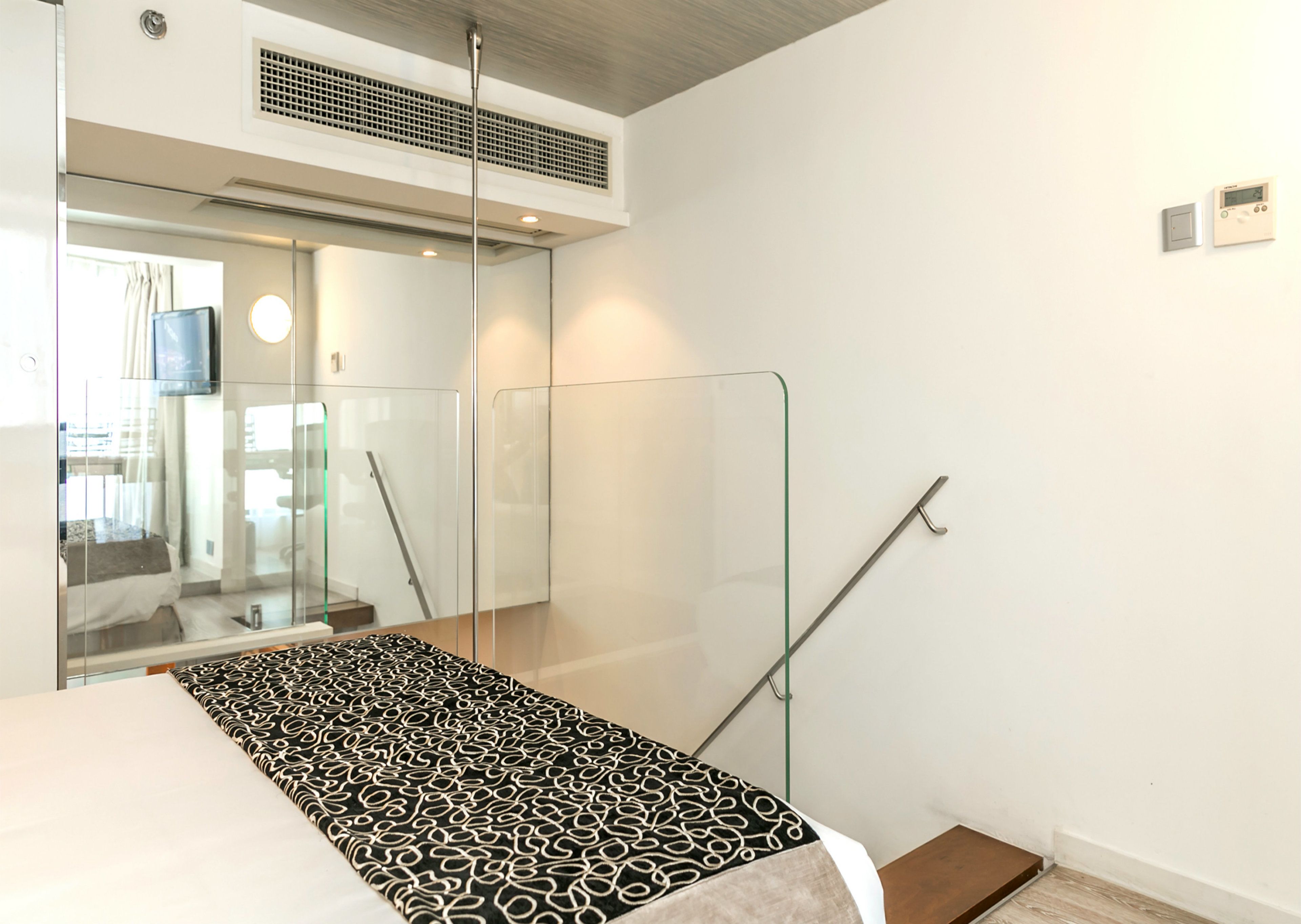 A modern hotel room with a glass partition, a bed with a patterned bedspread, a mounted thermostat, and a visible staircase.