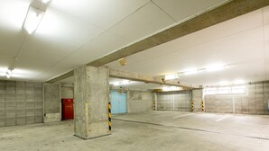 Uma ampla garagem subterrânea vazia, com paredes de concreto, um telefone vermelho de emergência e uma porta azul ao fundo.