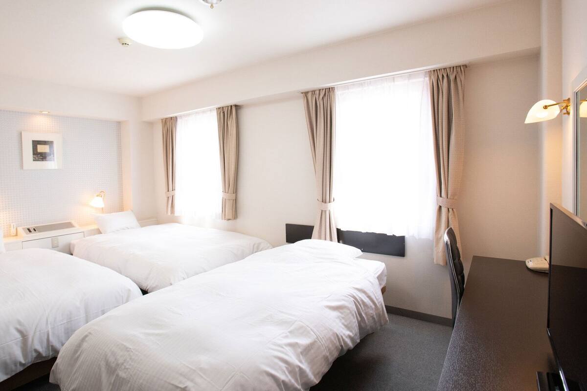 Habitación de hotel con dos camas, una mesita de noche con lámpara, un espejo y una ventana con cortinas.