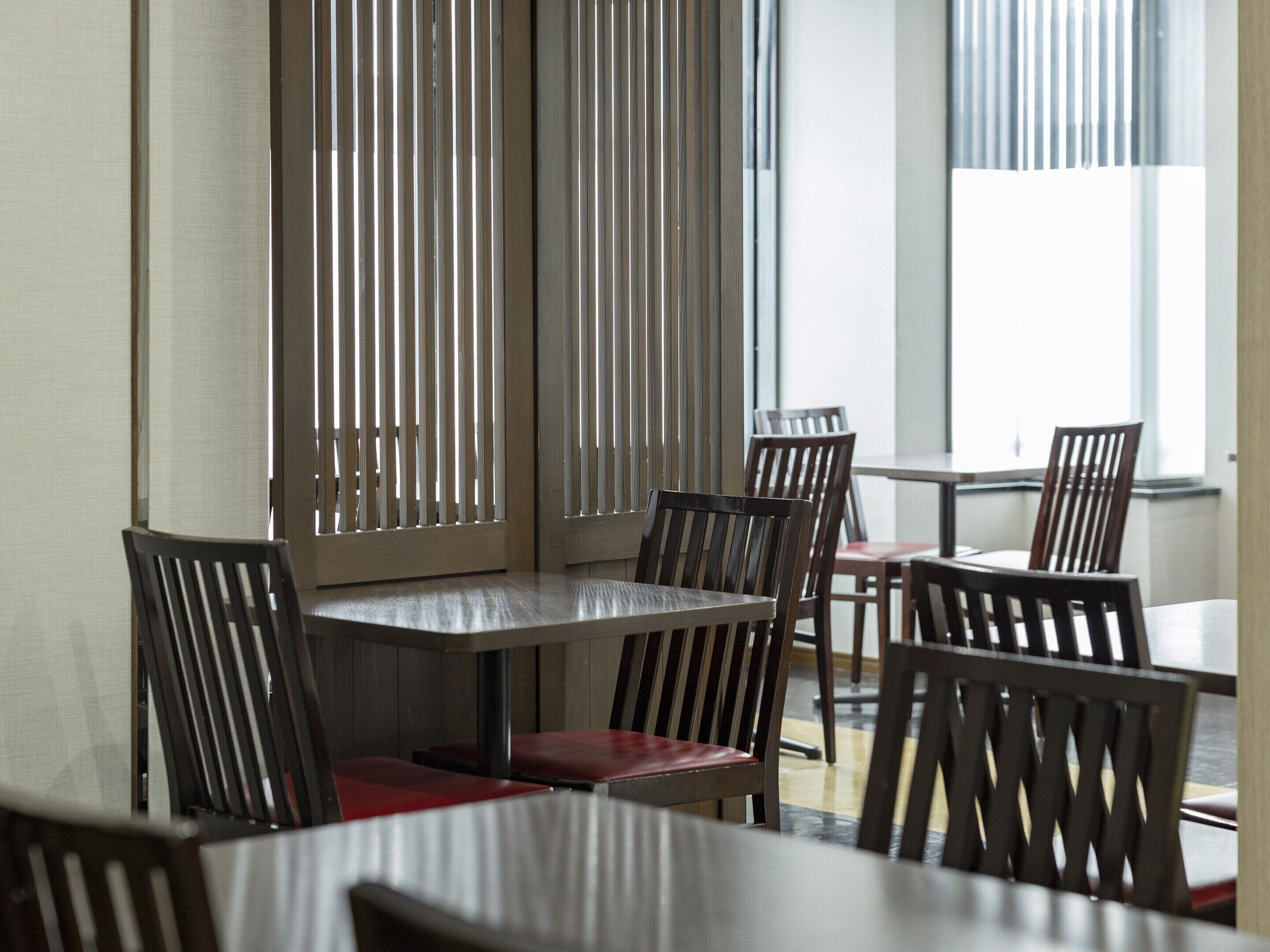 A restaurant interior with wooden tables and chairs, vertical blinds on the windows, and a view of the outside.