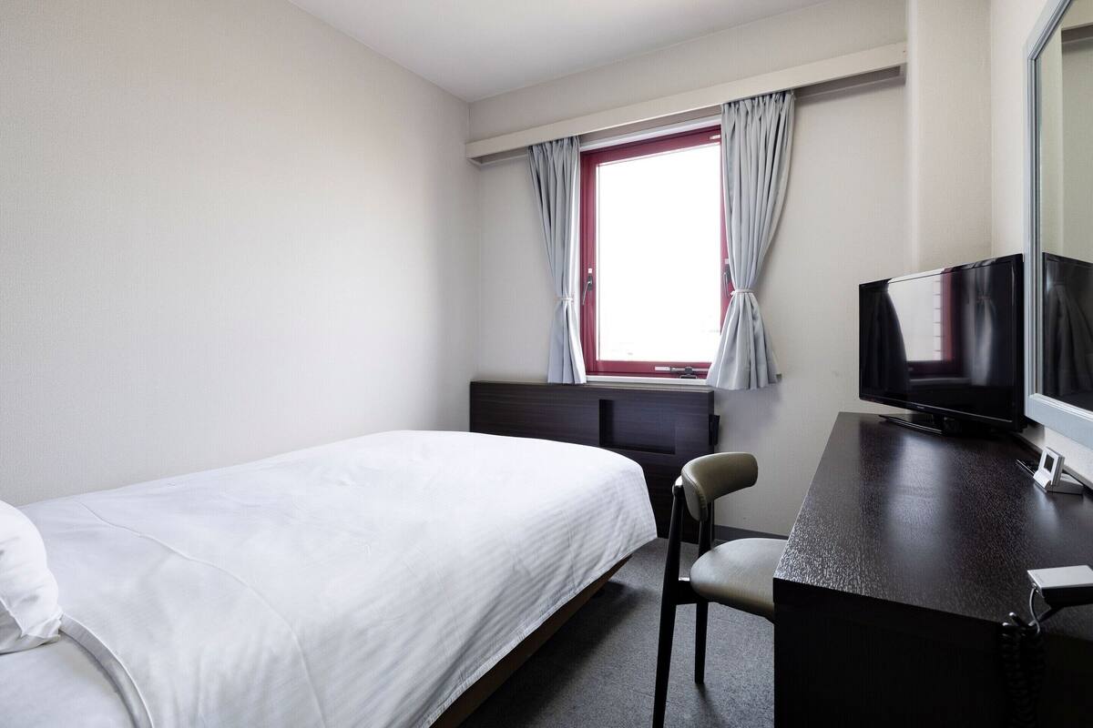 Habitación de hotel con una cama, un escritorio con televisor, una silla y una ventana con cortinas.