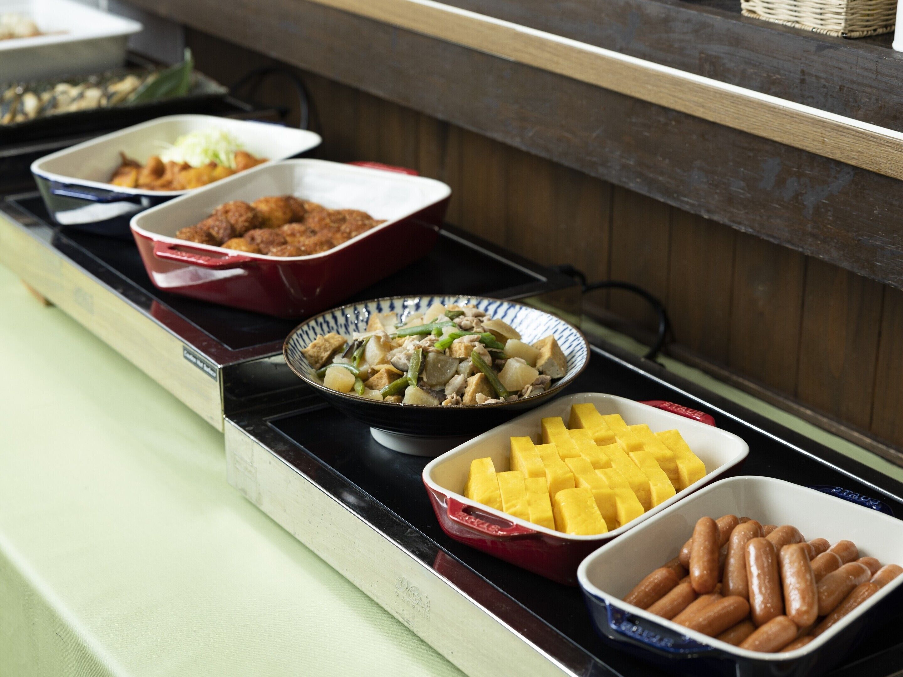 A buffet table with various dishes including a casserole, a stir-fry, mango chunks, and sausages.