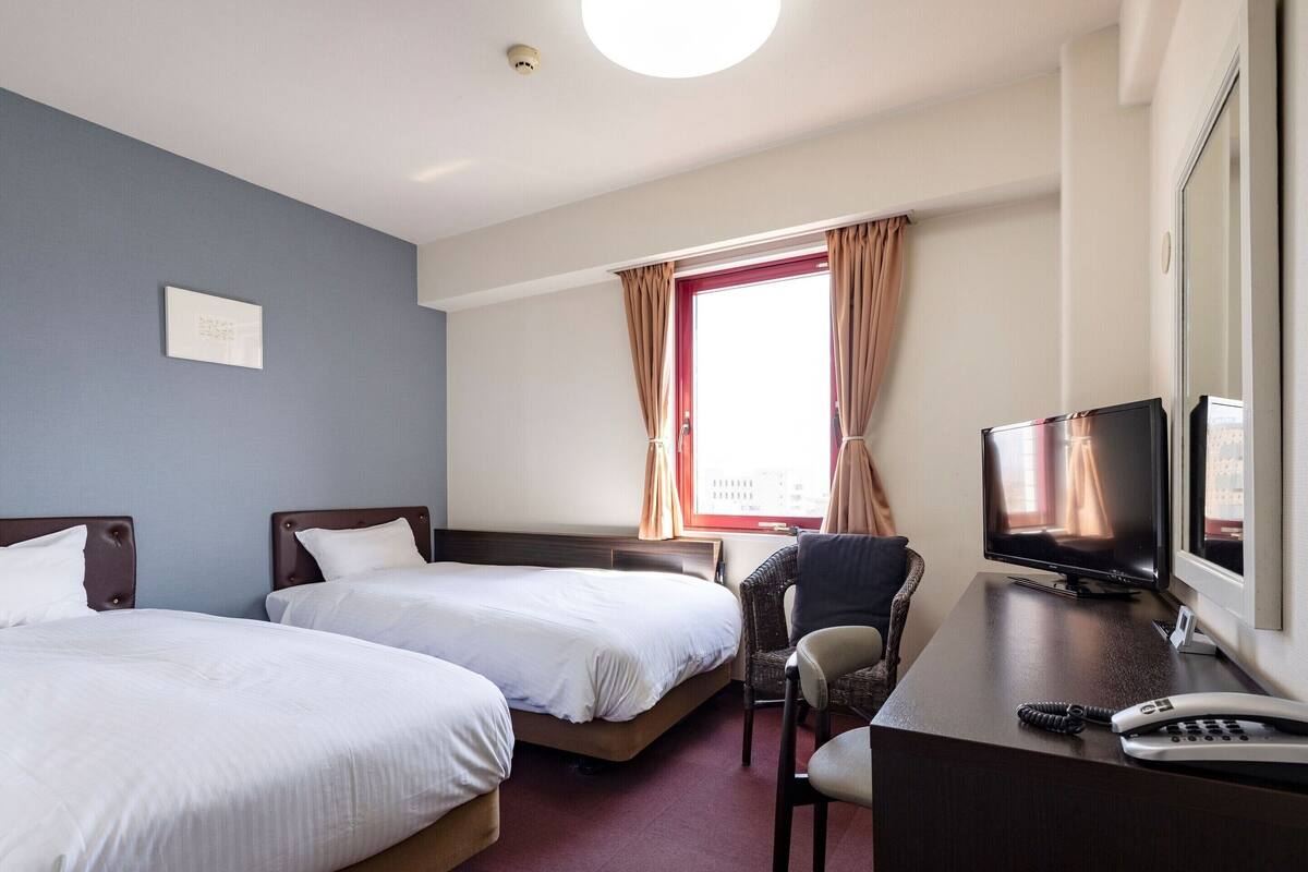 Habitación de hotel con dos camas, un escritorio con teléfono, una silla, televisor y ventana con cortinas.