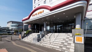 Entrada de hotel com um destaque para o letreiro 'KOKO STAY', uma escadaria que leva à entrada e uma placa de estacionamento.