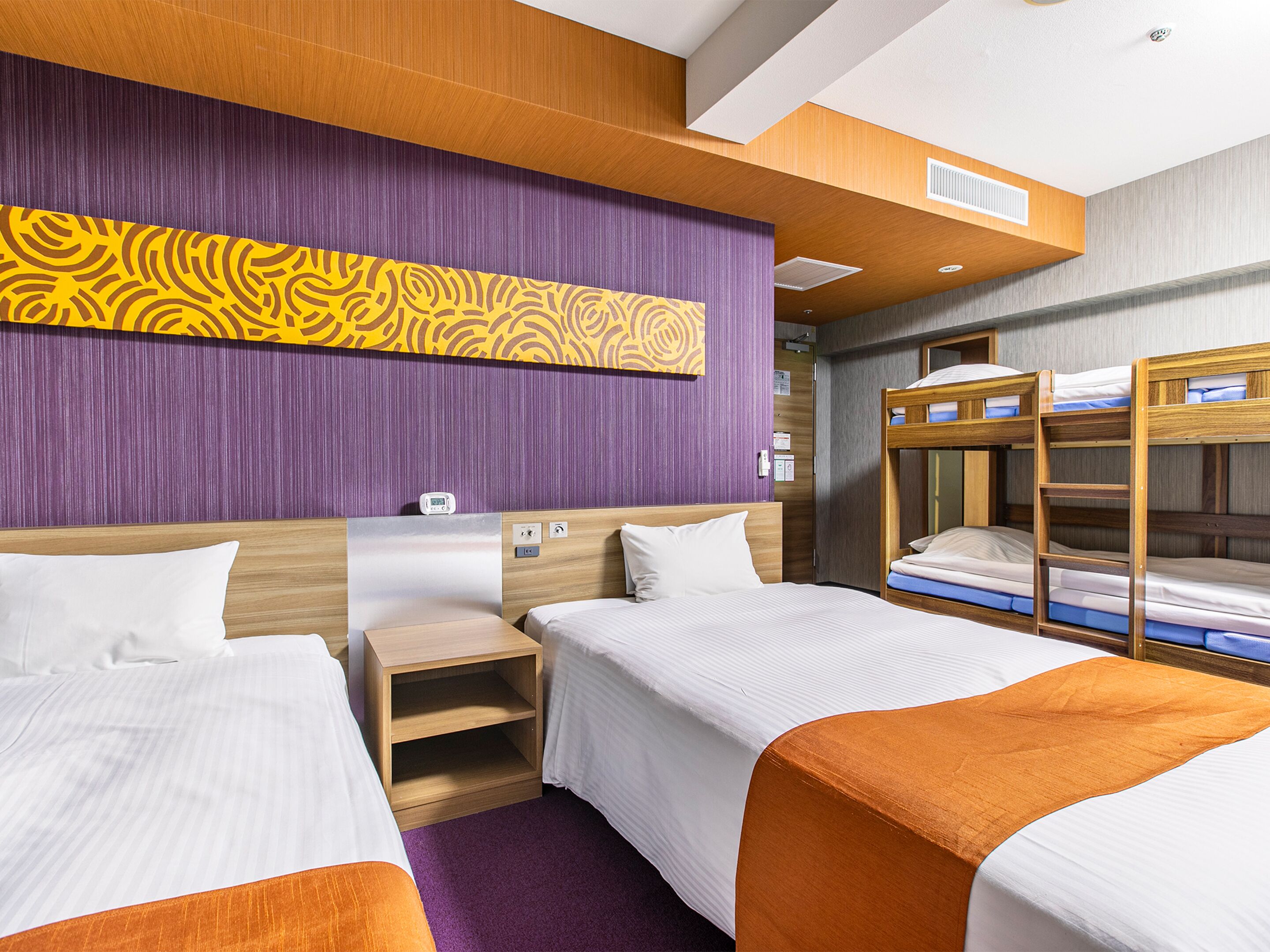 A hotel room with two single beds, a bunk bed, a wooden nightstand, and a purple accent wall.