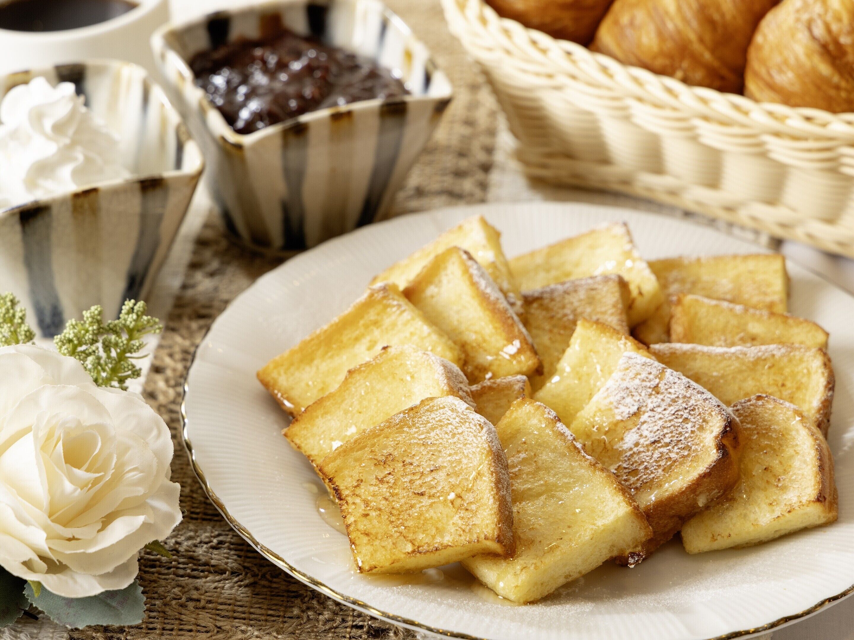 Uma fatia de pão francês com calda e açúcar em pó, uma rosa branca e uma cesta de croissants.