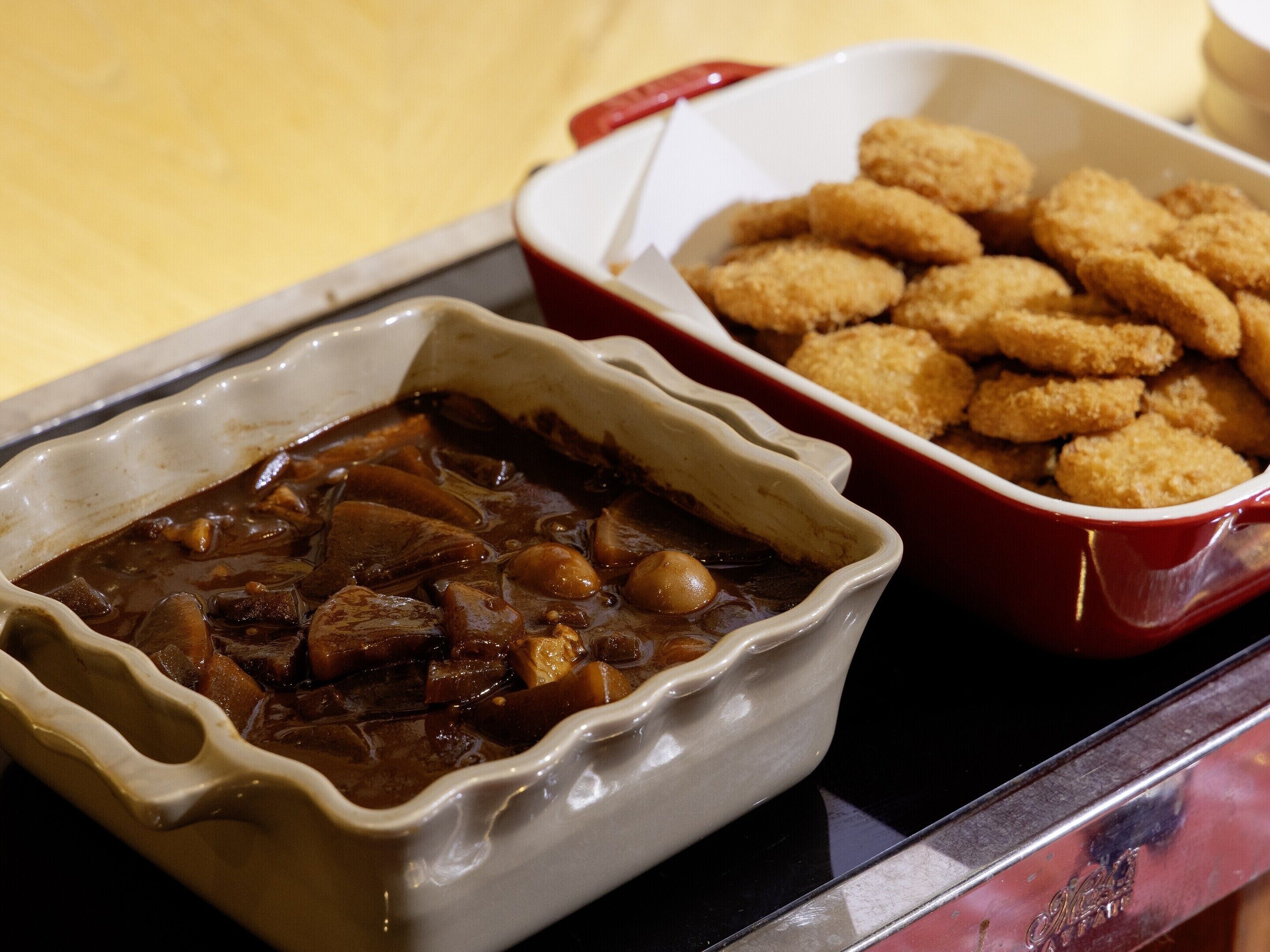 A dish of stew with chunks of meat and vegetables in a ceramic dish, accompanied by a side of breaded items in a red dish.