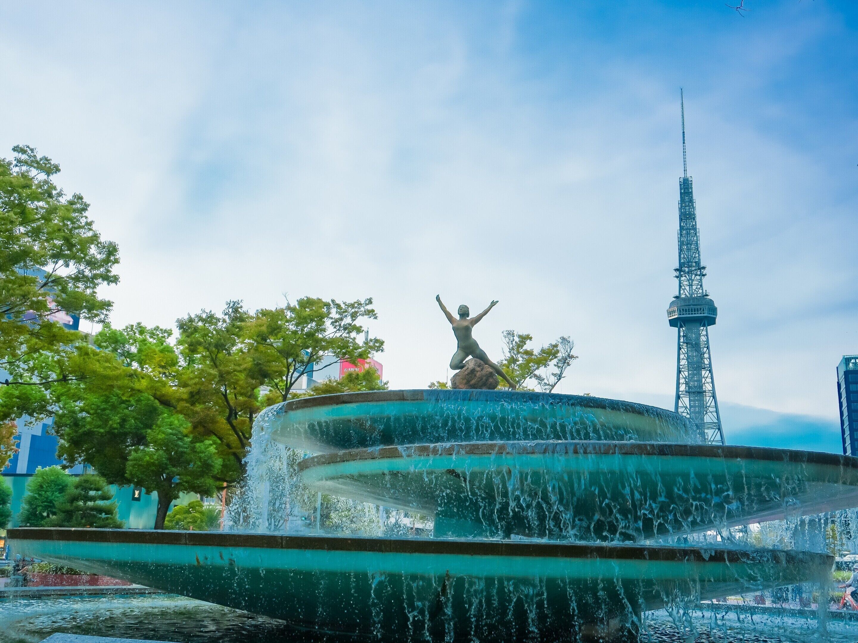 Une fontaine ornée d’une statue au centre, entourée d’arbres et avec une haute tour en arrière-plan.