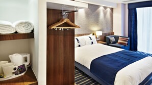 Ein Hotelzimmer mit einem Bett, einem Nachttisch mit Lampe, einem Sessel, einem Kleiderhaken und einem Regal mit Handtüchern und einer Kaffeemaschine.