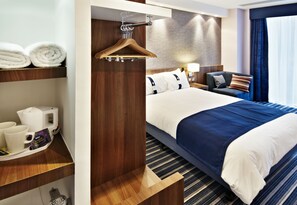Una habitación de hotel con cama, mesita de noche con lámpara, silla, perchero y estante con toallas y cafetera.
