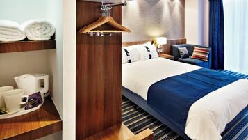 Una habitación de hotel con una cama, una mesita de noche con lámpara, una silla, un perchero y un estante con toallas y cafetera.