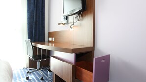 Ein kleines Hotelzimmer mit einem Holztisch, einem Stuhl, einem an der Wand montierten Fernseher und einer kleinen, violetten Ablage.
