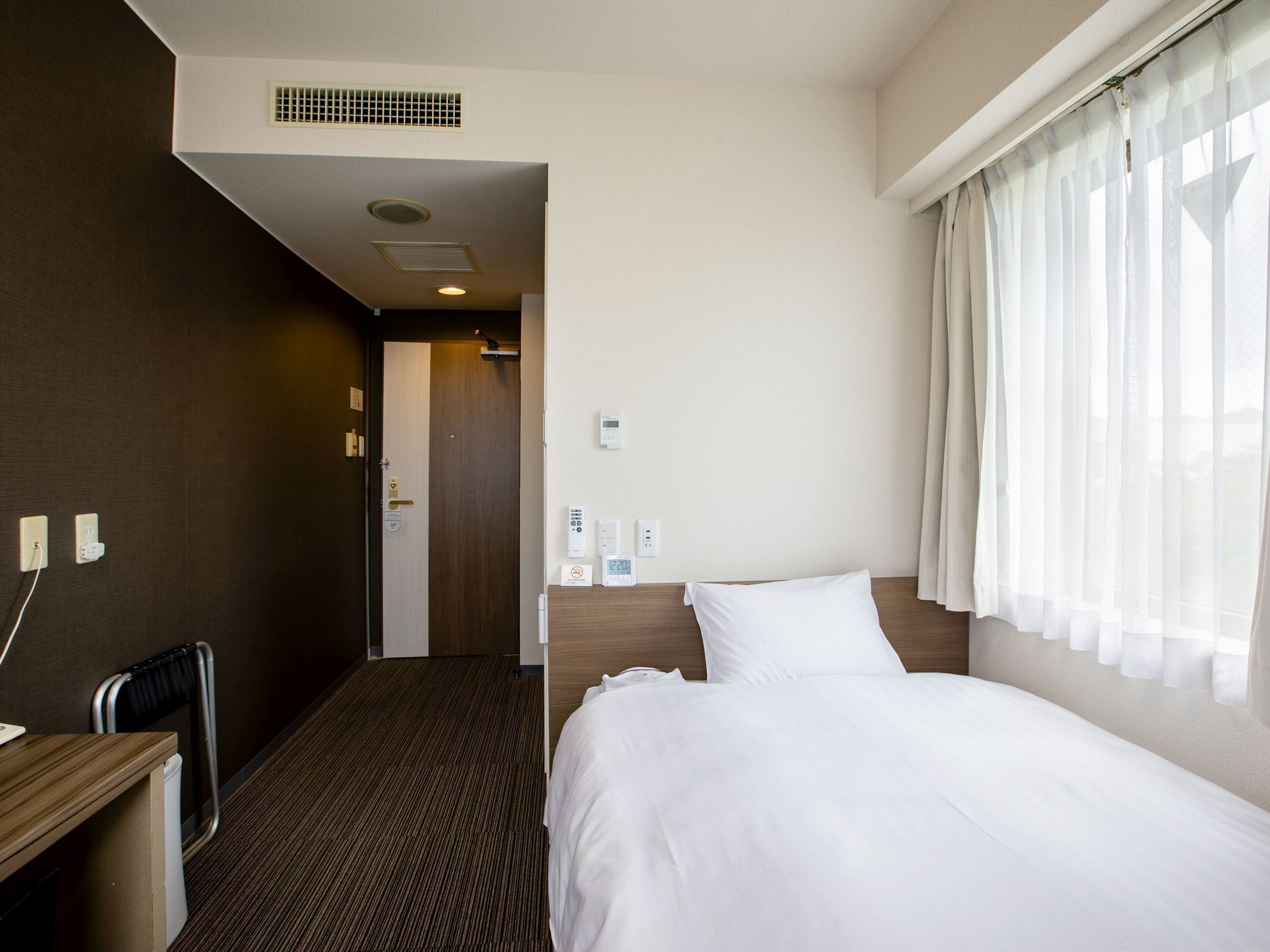 Une chambre d’hôtel comprenant un lit, un bureau, une chaise, un radiateur, une fenêtre avec des rideaux et une porte donnant sur une autre pièce.