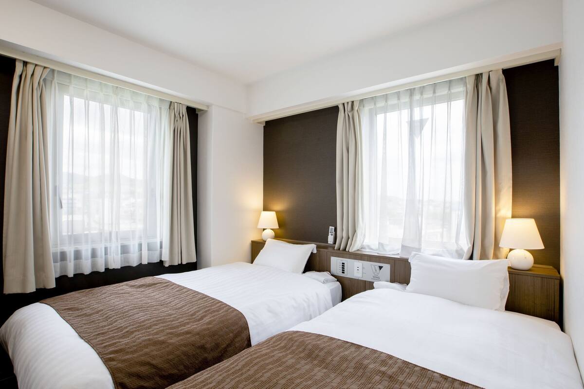 A hotel room with two beds, a bedside table with a lamp, a control panel, and sheer curtains.