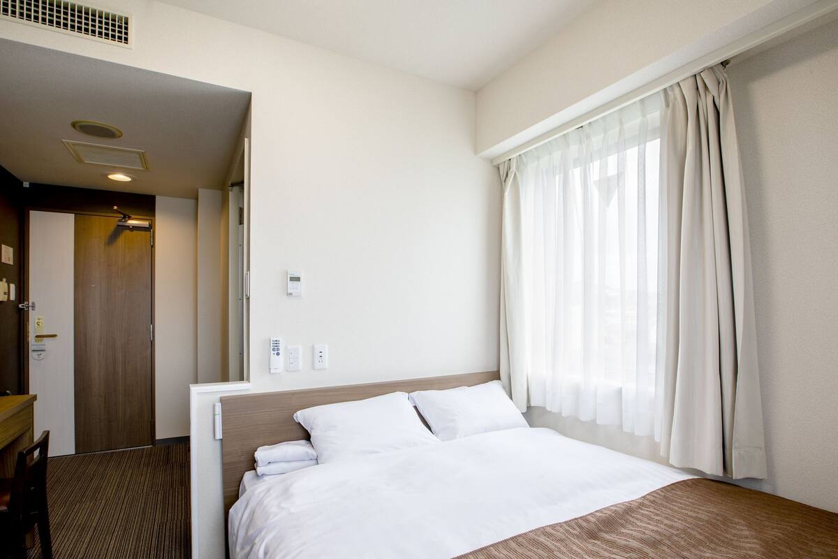 A hotel room with a large bed, a wooden headboard, a desk with a chair, and a window with sheer curtains.