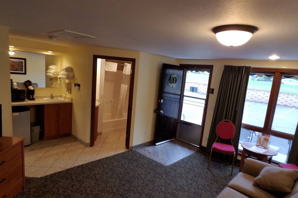 A hotel room with a kitchenette, a bathroom with a shower, a living area with a sofa, and a view of the outside.