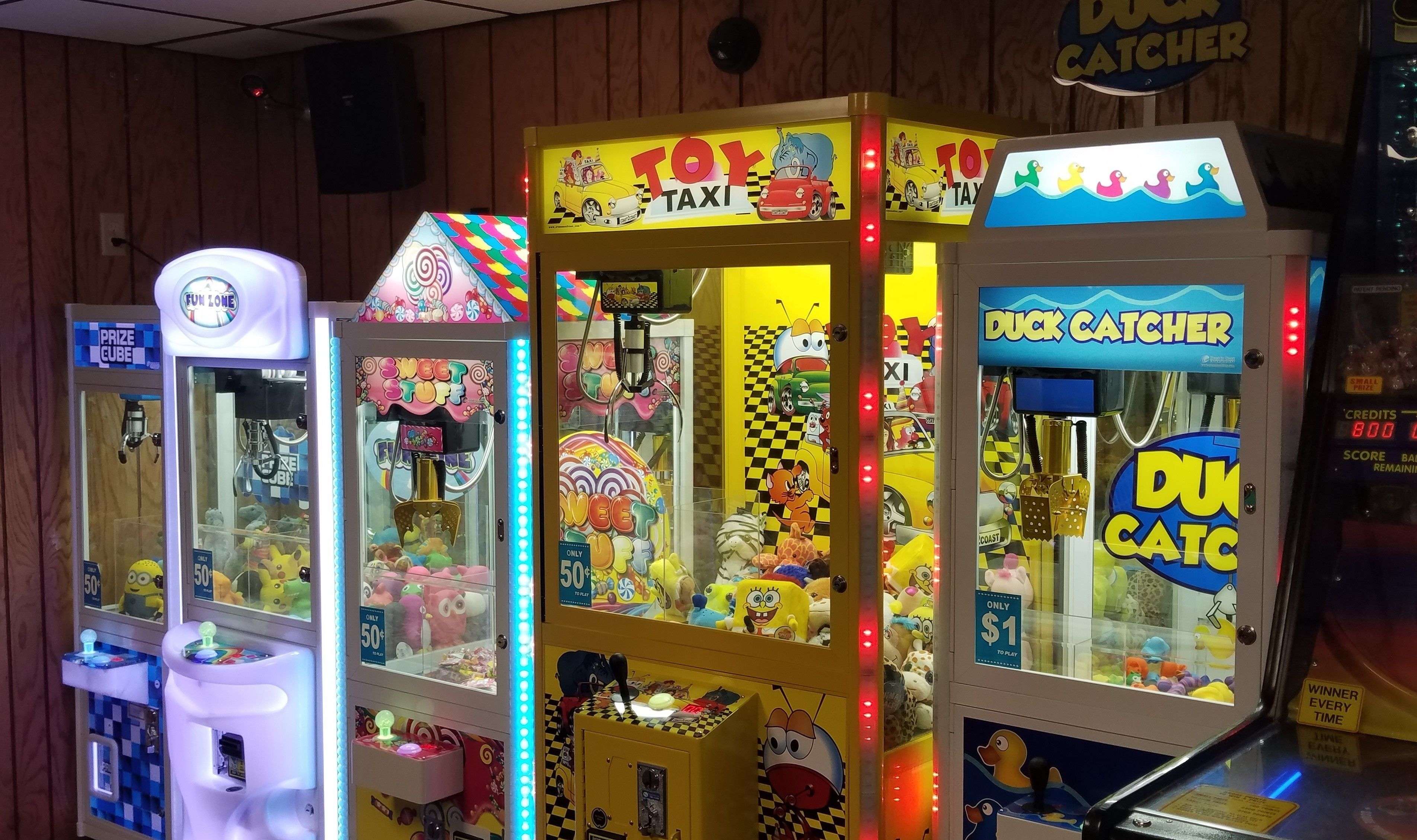 A row of brightly lit claw machines with various themes, including a toy taxi and a duck catcher.