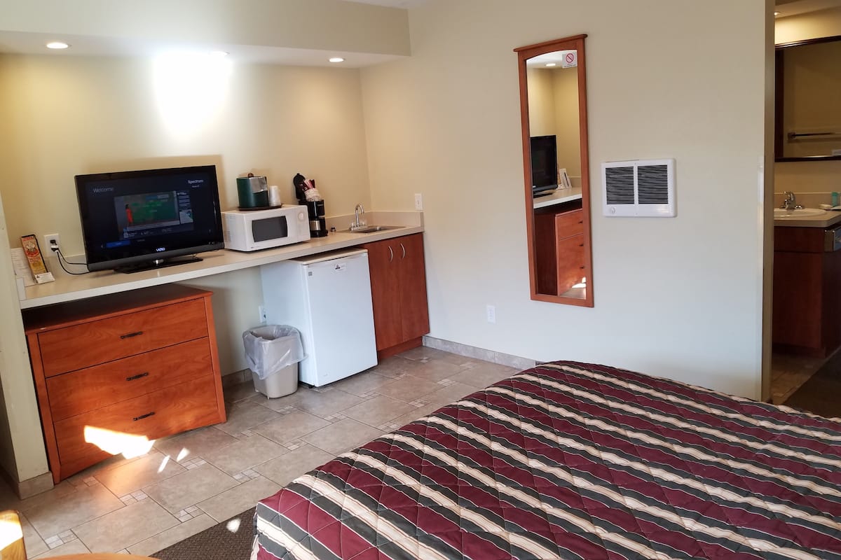 A compact hotel room with a bed, a small kitchenette, a television, and a mirror.