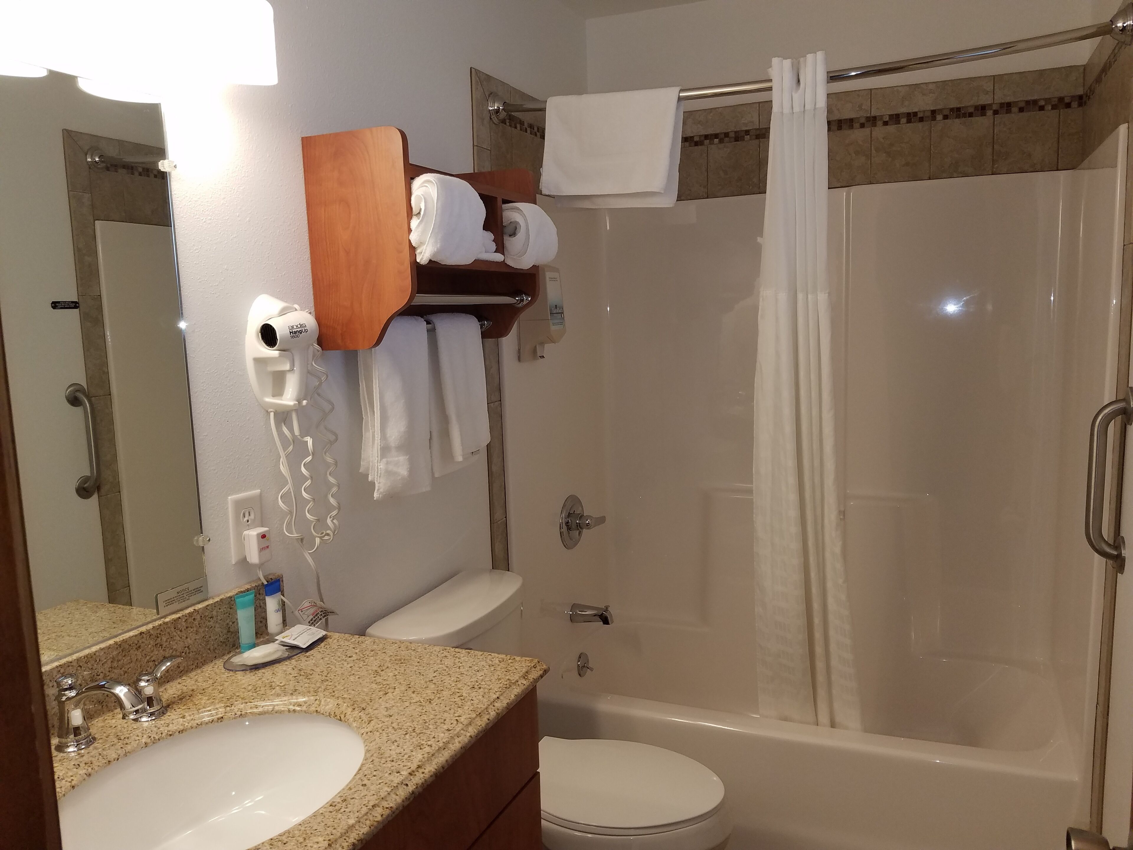 Un baño con lavamanos, inodoro y ducha. Se ve un perchero con toallas y un secador de pelo.