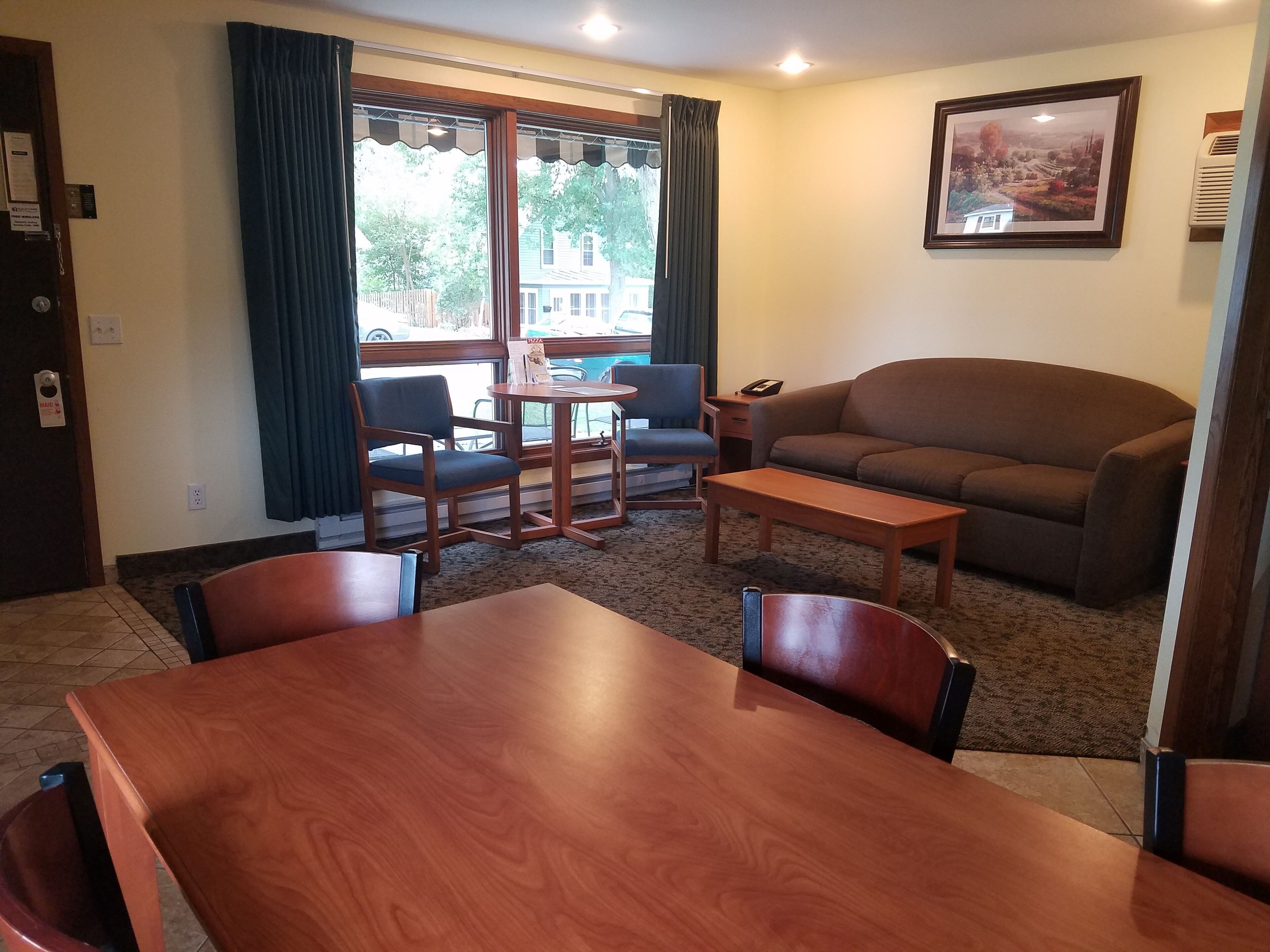 A living room with a brown sofa, two armchairs, a coffee table, and a dining table. There is a window with curtains, a framed picture on the wall, and a door with a sign.