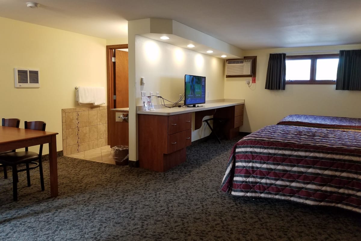 A hotel room with a bed, a desk with a computer, a chair, a small table, a bathroom with a toilet, and a window with curtains.