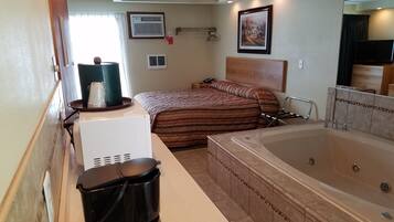 A compact hotel room with a bed, a small desk, a coffee maker, a large bathtub, and a mirror.