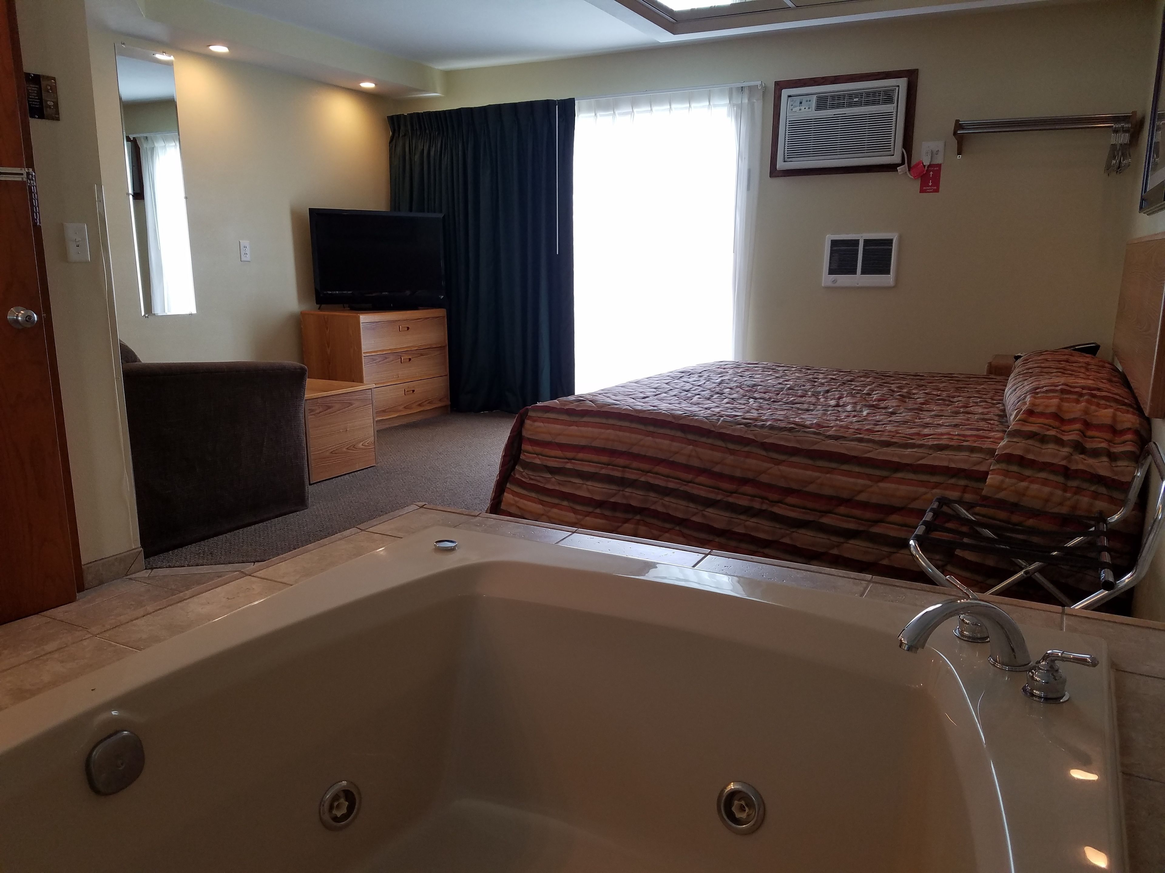 Ein Hotelzimmer mit einer großen Badewanne, einem Bett mit gestreifter Bettwäsche, einem Fernseher, einem Schminktisch und einem Fenster mit Vorhängen.