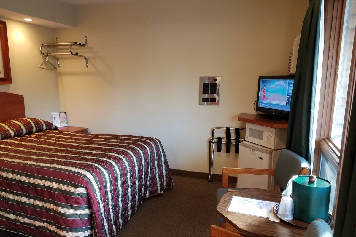 A hotel room with a bed, a desk with a computer, a microwave, and a small table with a lamp.