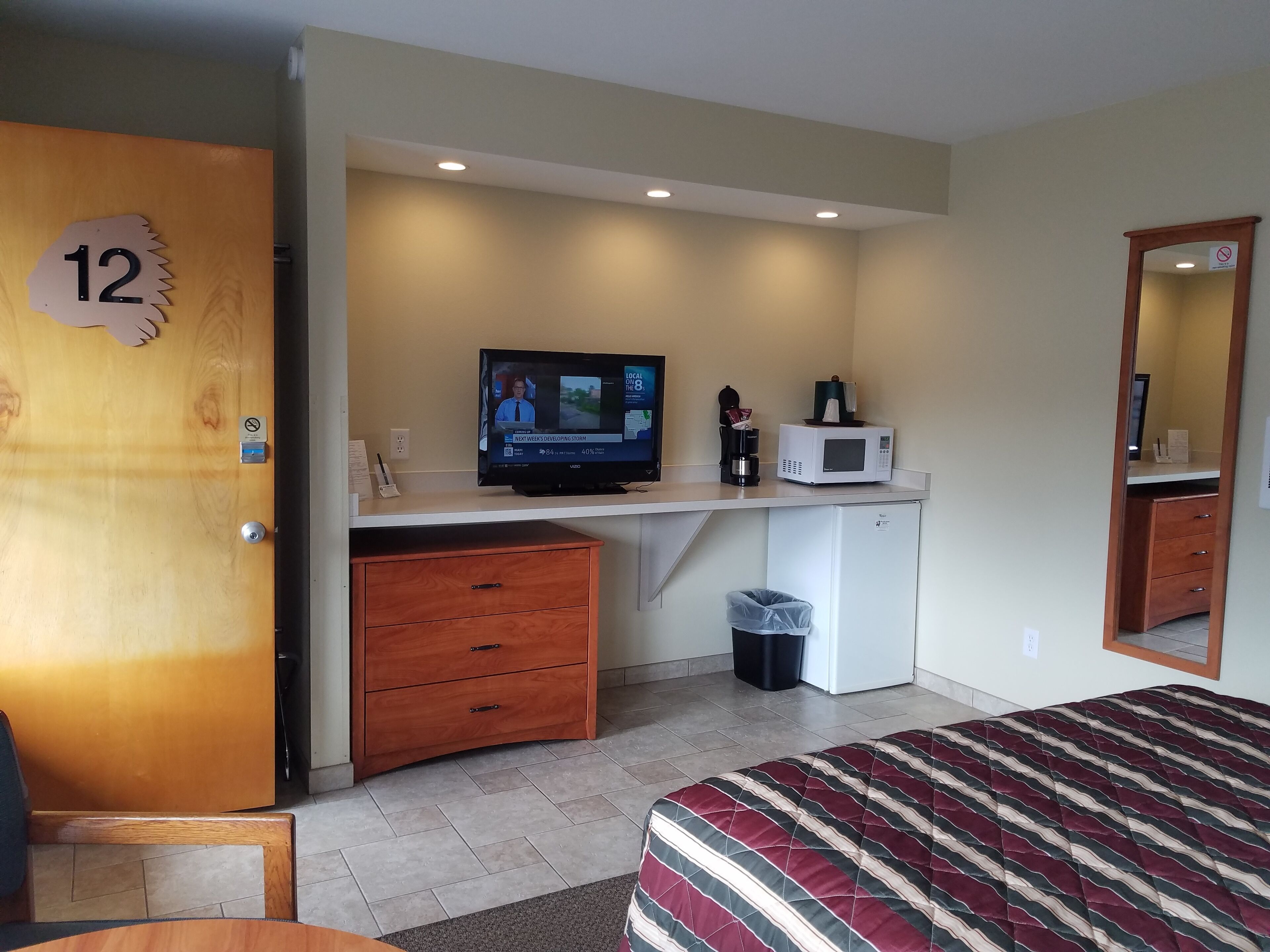 A hotel room with a wooden desk, a television, a microwave, a trash bin, and a bed with a striped bedspread.