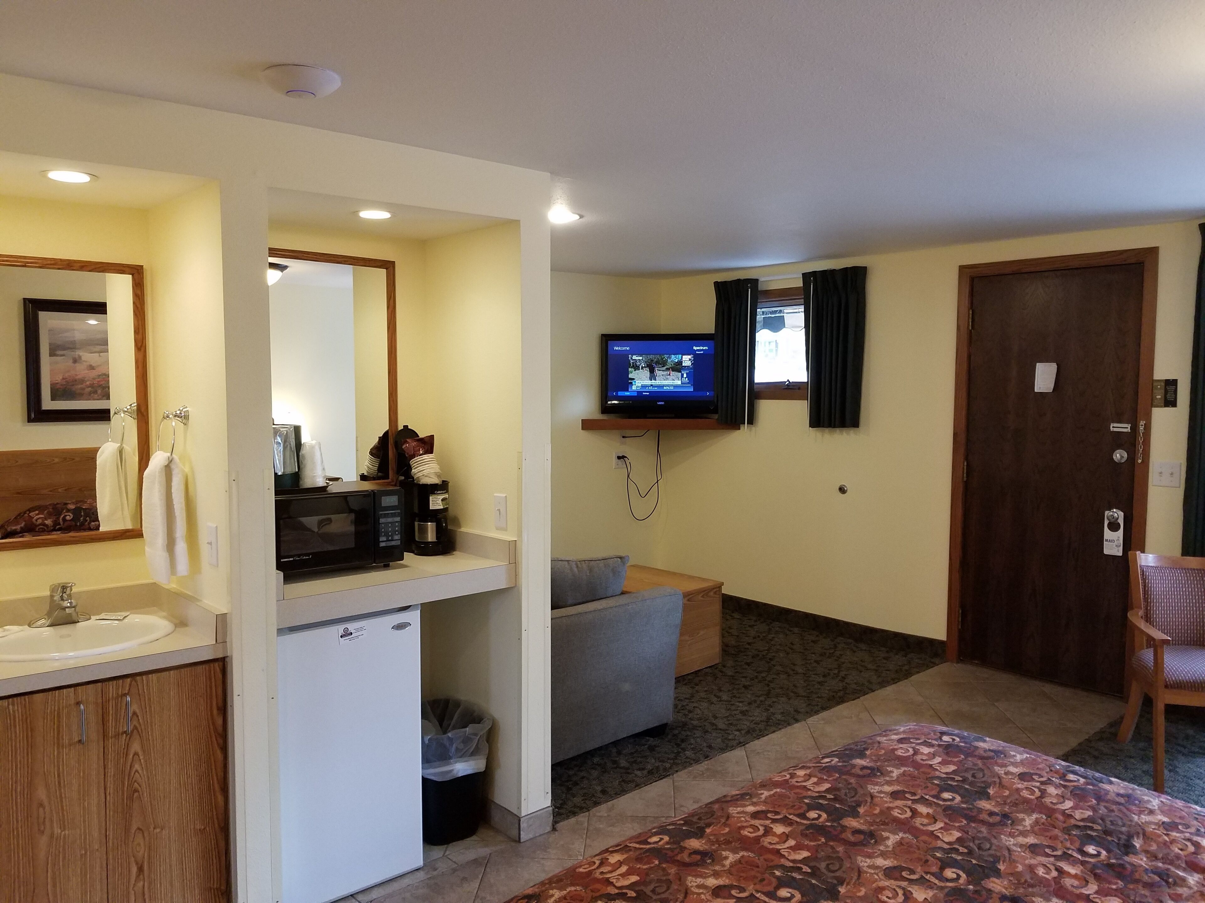 Ein Hotelzimmer mit Bad, einem an der Wand befestigten Fernseher, einer Couch und einer geblümten Bettdecke.