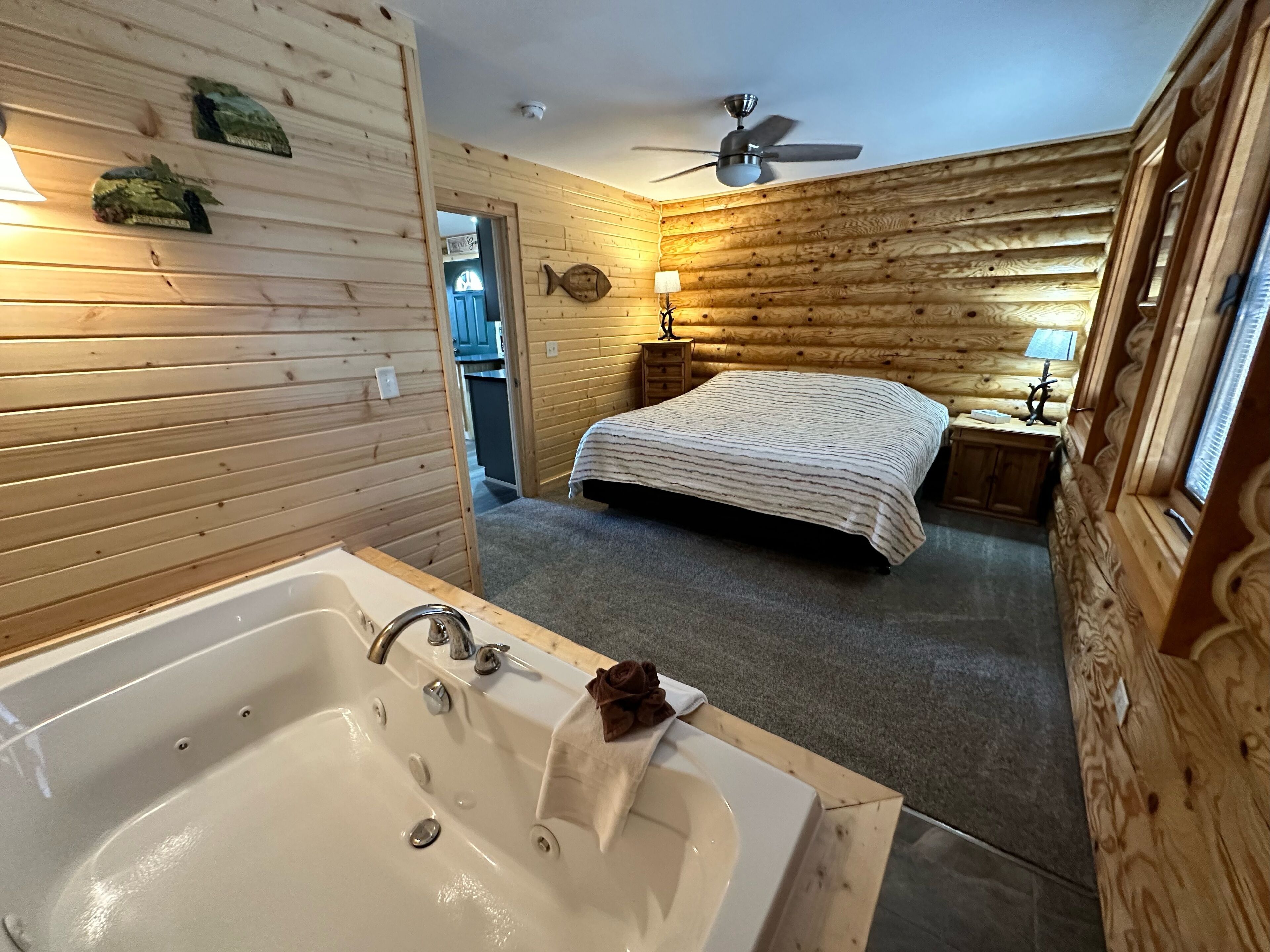 A wooden cabin bedroom with a large bathtub, a bed with a striped blanket, and a ceiling fan.