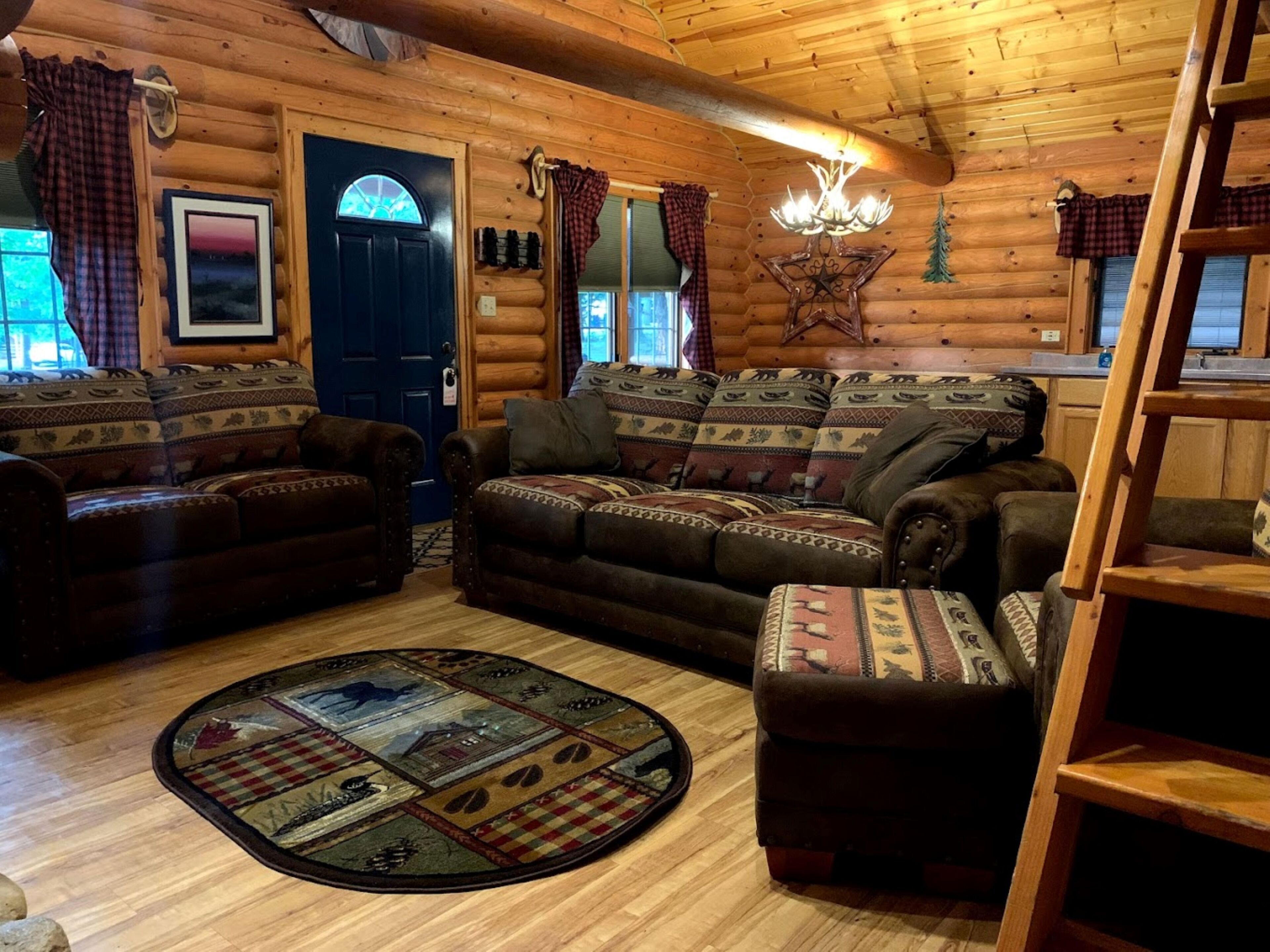 A cozy log cabin interior with a large sofa set, a round rug with a patchwork design, and a wooden ceiling.