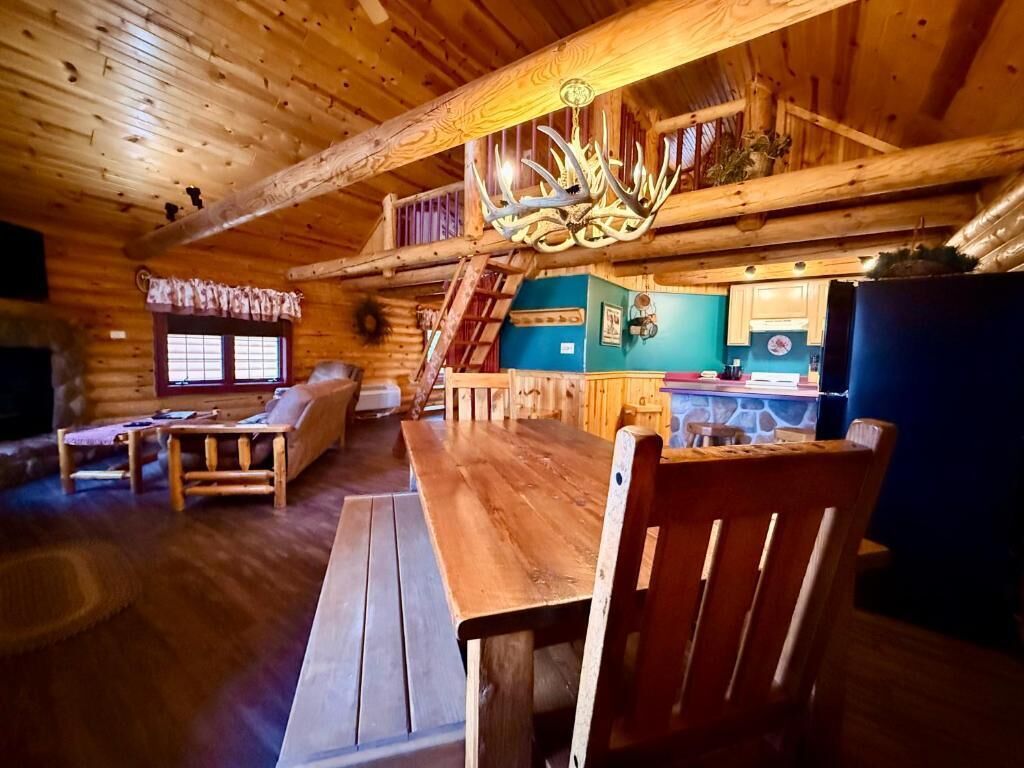 A rustic cabin interior with a wooden dining table, a sofa, and a staircase leading to an upper level.
