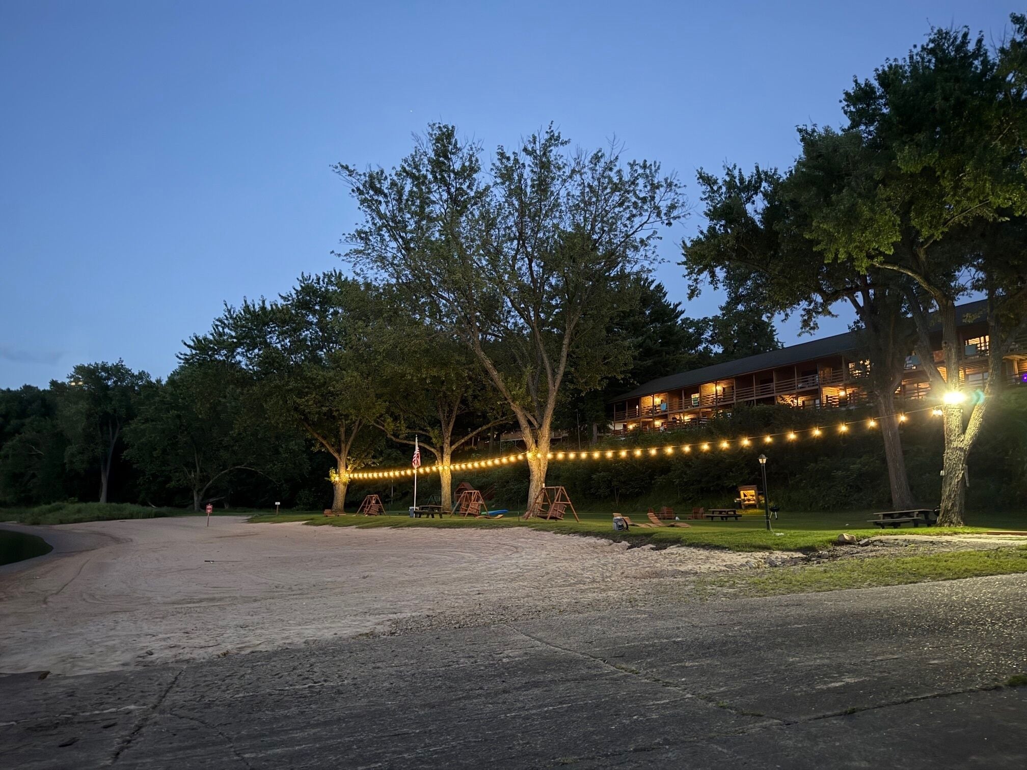 A lodge with a large deck and string lights, surrounded by trees and a gravel path.