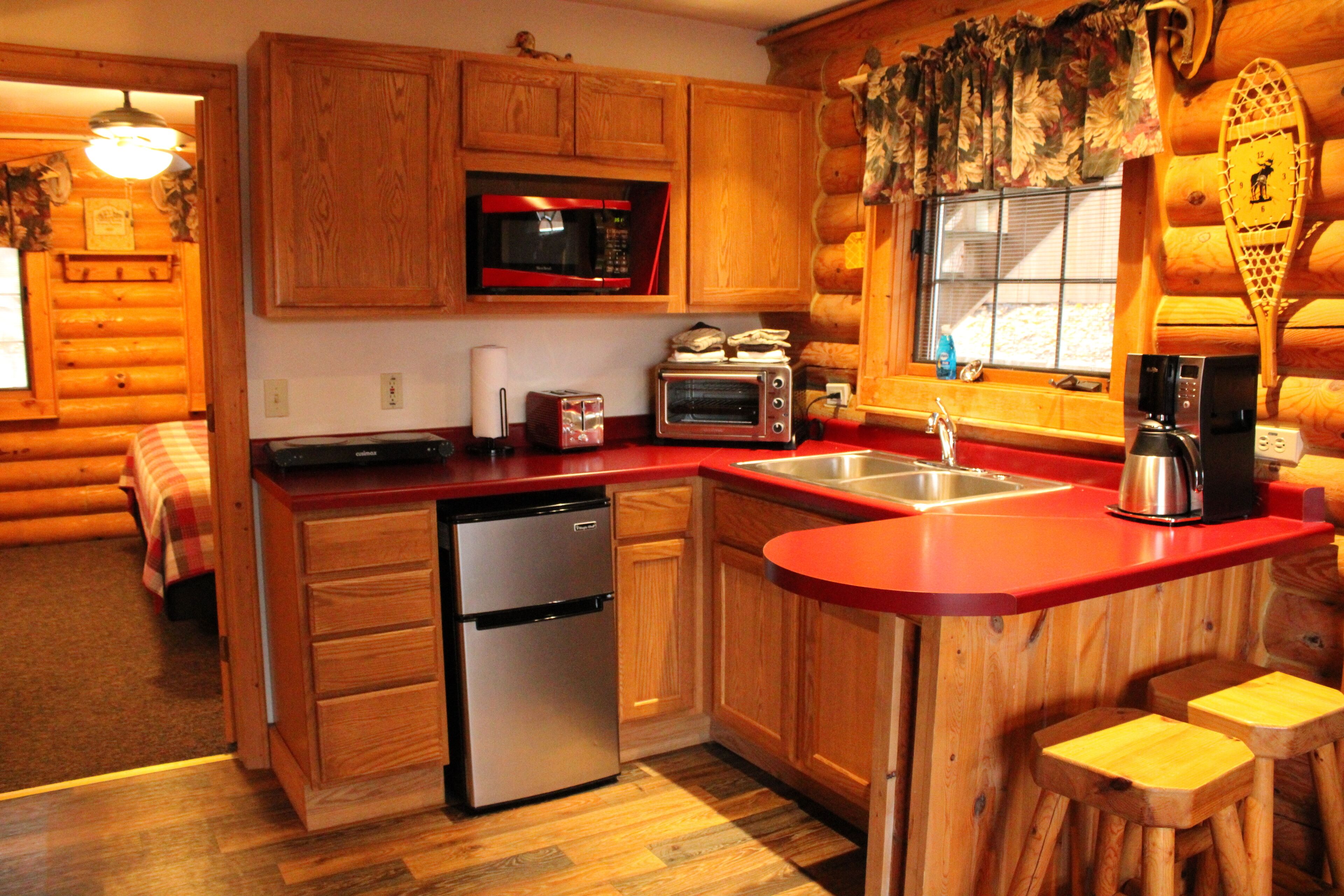 A log cabin-style kitchen with wooden cabinets, a stainless steel dishwasher, a red countertop, and a window with a decorative valance.