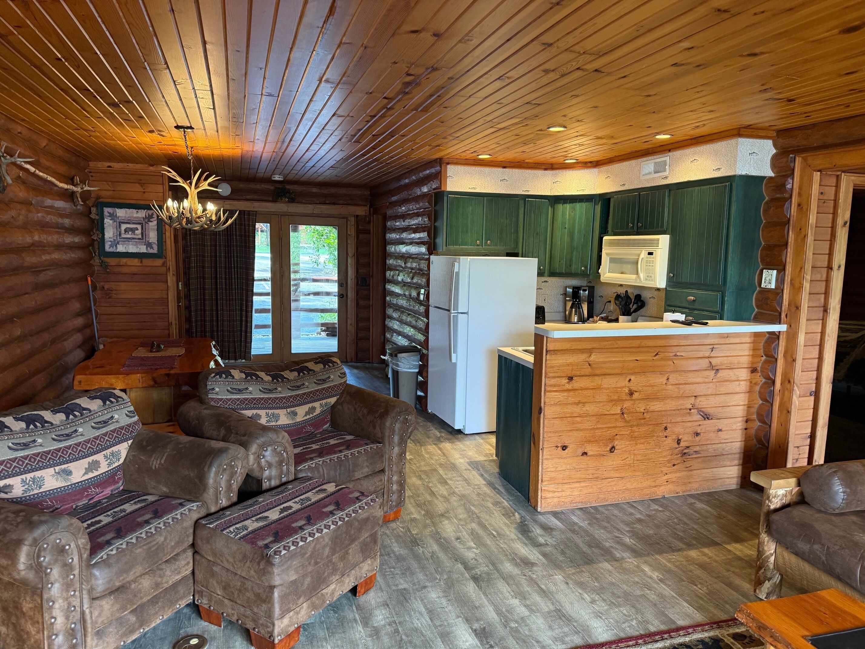 A log cabin interior with a living area featuring leather sofas and a kitchen with green cabinets and a white refrigerator.