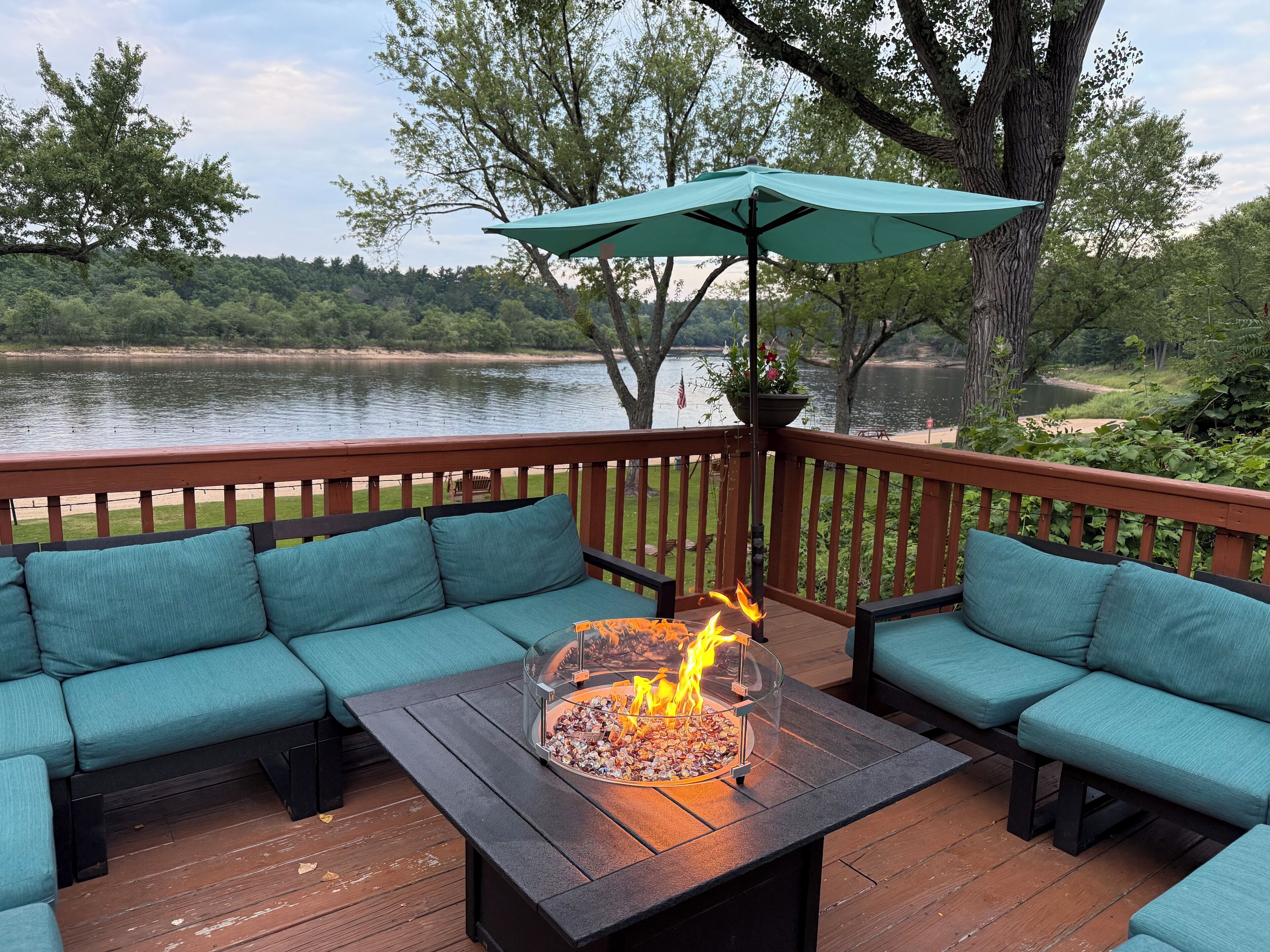 Une terrasse dotée d’un foyer intégré, de sièges confortables aux coussins turquoise et offrant une vue sur un lac.