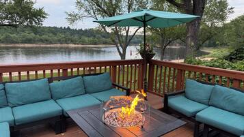 Une terrasse dotée d’un foyer intégré, de sièges confortables aux coussins turquoise et offrant une vue sur un lac.
