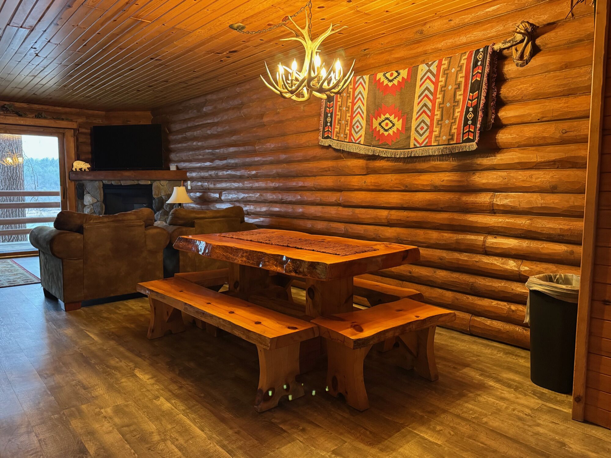 A log cabin interior with a wooden dining table, benches, a sofa, a fireplace, and a chandelier.