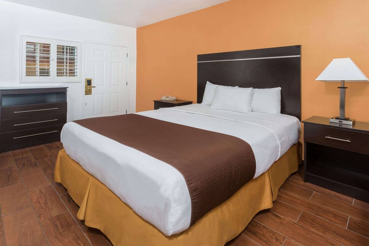 A hotel room with a large bed, a nightstand, a lamp, a dresser, and a window with blinds.