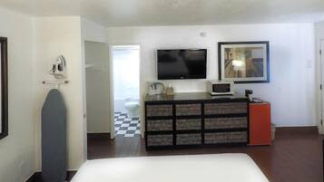 A hotel room with a bed, a TV, a small desk with a mirror, a dresser, and a door leading to a bathroom with a checkered floor.