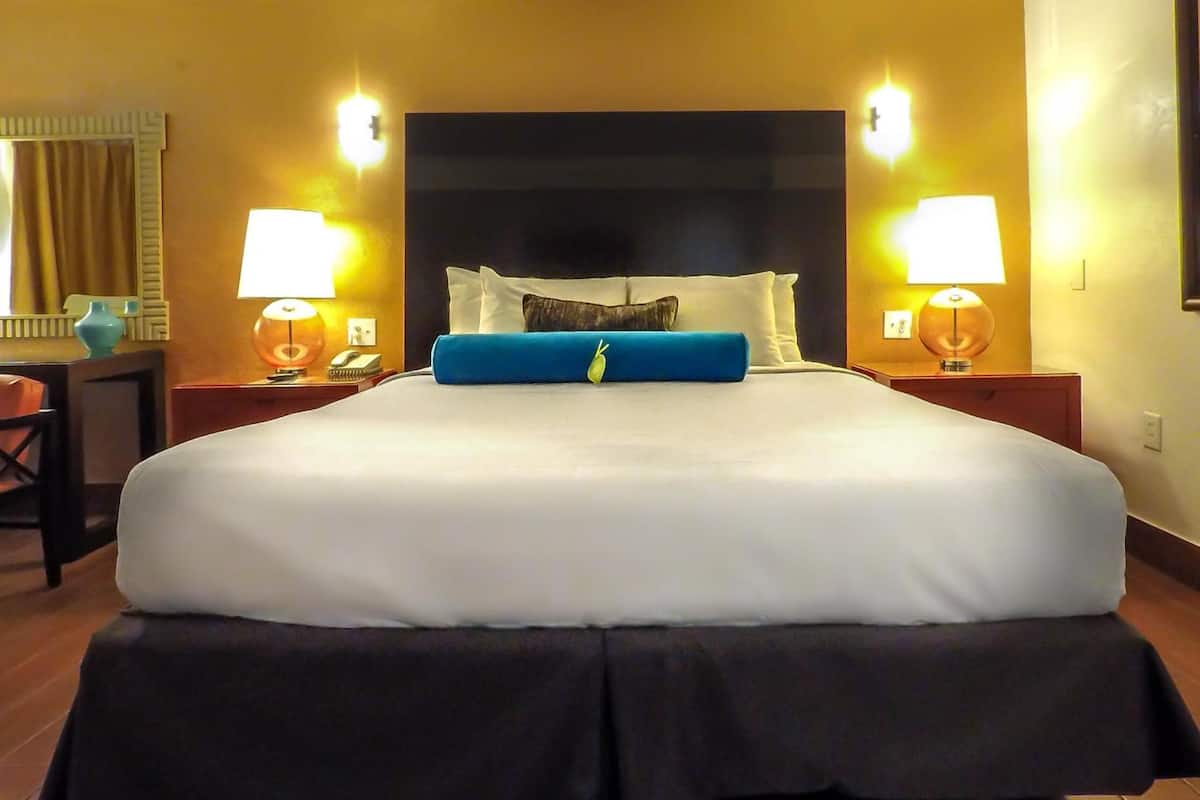 A hotel room with a large bed, two bedside lamps, a nightstand with a phone and a vase, and a mirror on the wall.