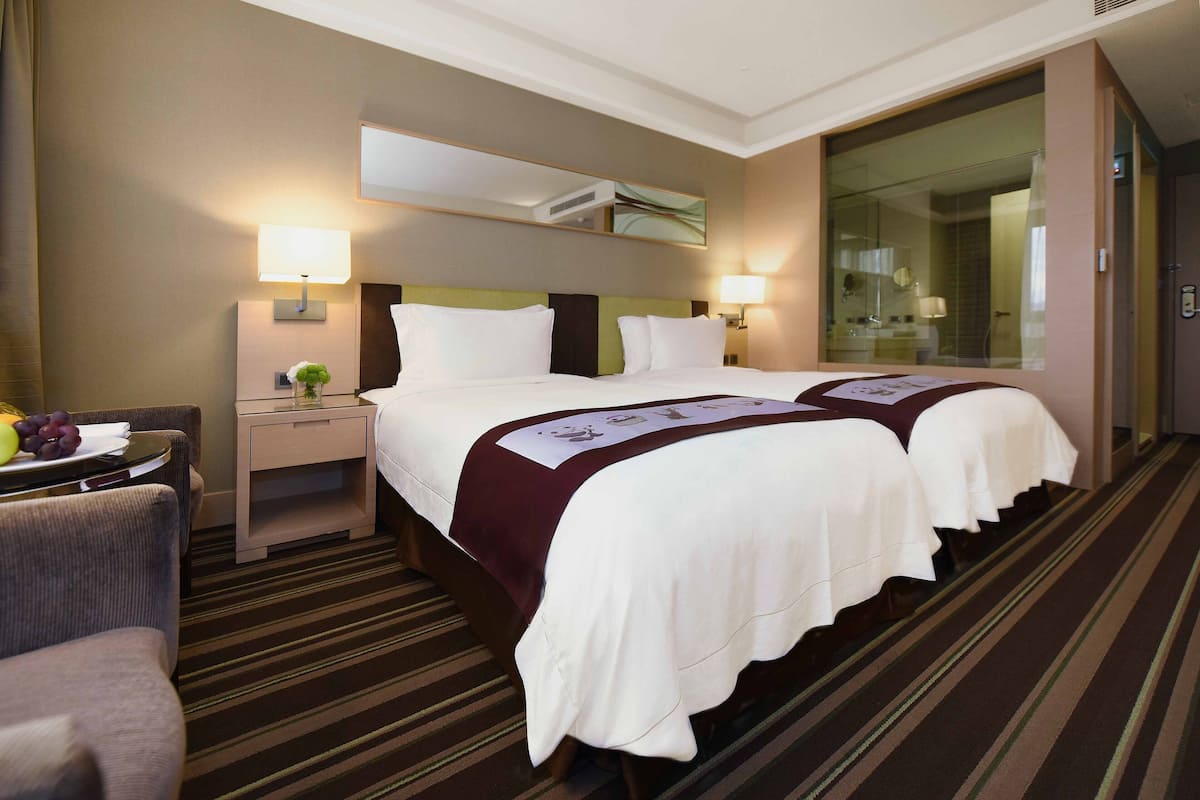 Ein Hotelzimmer mit zwei Betten, einer Sitzecke mit einem Sessel, einem kleinen Tisch mit Obst und einem großen Fenster.