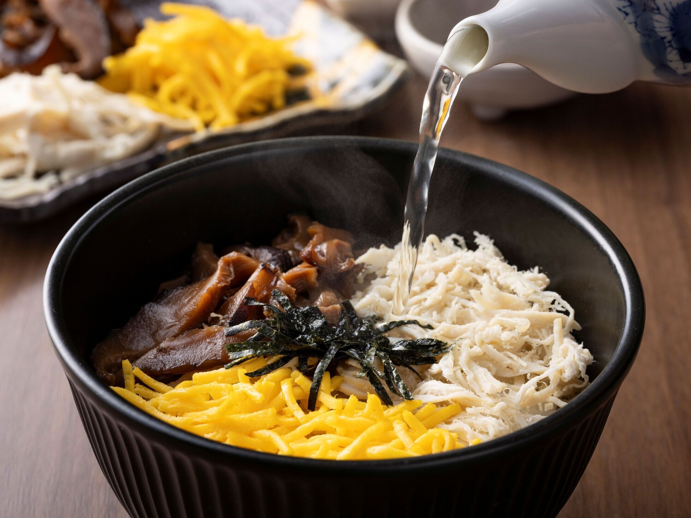 A bowl of food with various ingredients, including shredded cheese, seaweed, and meat, being poured into it.